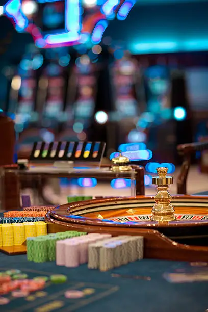 a focused view of a casino roulette table with a stack of chips on the spinning wheel and blurred gaming chips in the foreground. The background is softly out of focus, showing the bright, colorful lights typical of a casino environment, giving a sense of depth and the bustling atmosphere of the gaming floor.