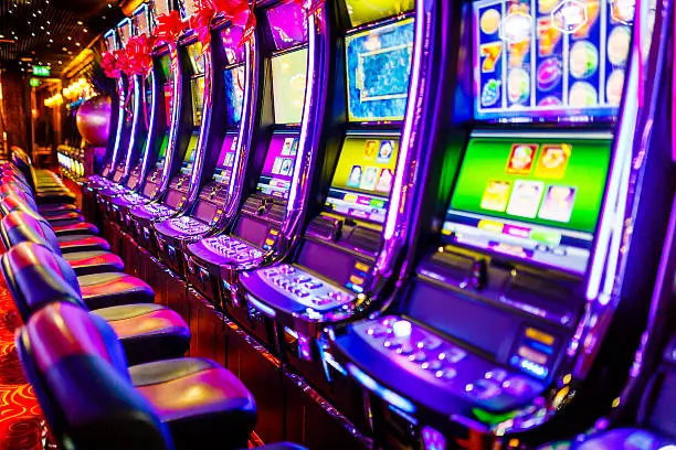a row of vibrant, illuminated slot machines at a casino, with no players present, suggesting either anticipation for the next round of play or a quiet moment in the gaming space. The colorful screens and flashy lights create an atmosphere of excitement and entertainment, while plush chairs in front wait for players to take a seat.